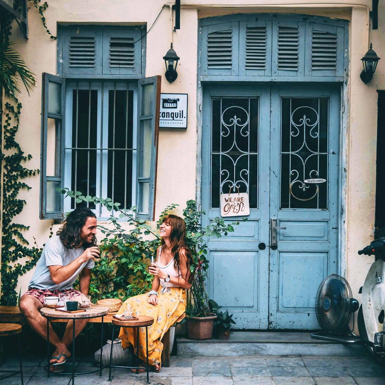 Couple sitting at cafe table outside blue door with shuttered windows, reading and enjoying coffee