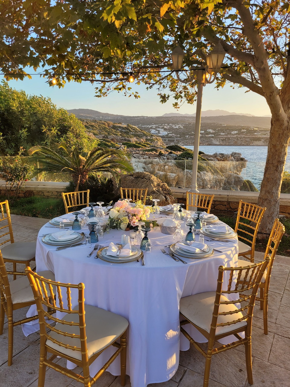 Round dining table with white linens and floral centerpiece on terrace overlooking Rethymno harbor and sea at sunset