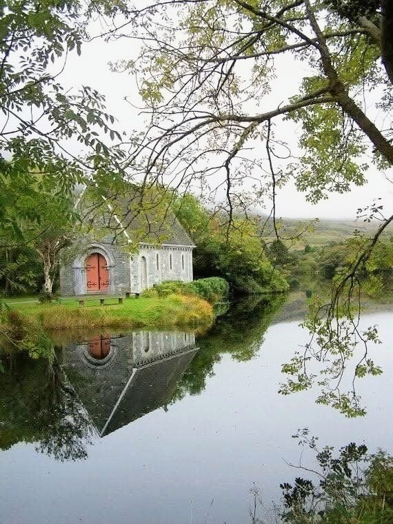 White church with red door beside tranquil lake surrounded by trees and green foliage with mirror-like reflections