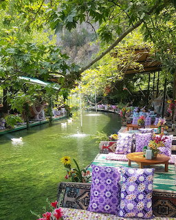 Outdoor riverside café with colorful cushions and rugs beside a green stream surrounded by lush vegetation