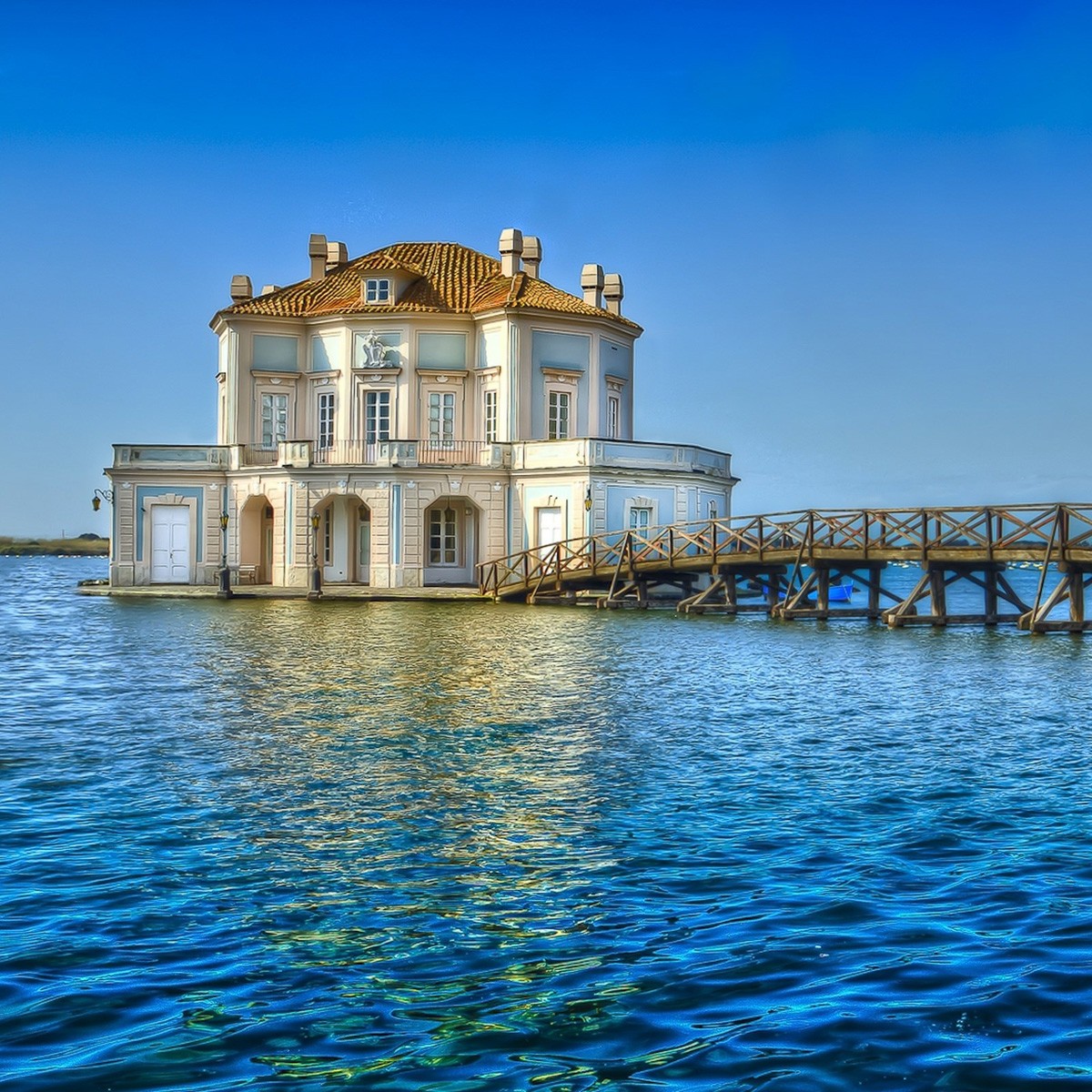 White villa with yellow roof standing in blue water, connected by wooden bridge