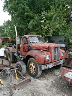 Rusty red vintage Mack truck in a junkyard surrounded by scrap and trees