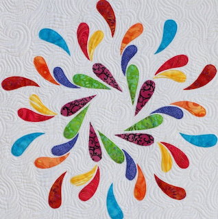 Colorful quilt block with teardrop shapes in rainbow colors arranged in circular splash pattern on white quilted fabric