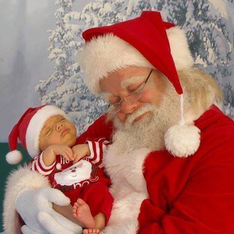 Santa Claus holding a sleeping newborn baby in a red Christmas outfit