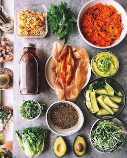 Flat lay of healthy foods including avocado, sprouts, vegetables, pasta, and a smoothie bottle on grey background