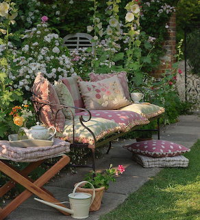 Iron garden bench with colorful cushions surrounded by flowers and a tea set on a folding table