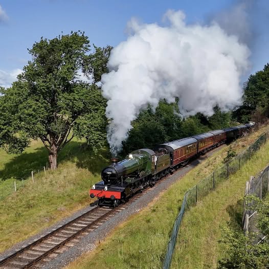Steam train with white smoke moving through rural landscape with trees and grass embankments