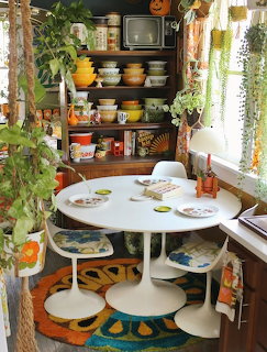 Retro kitchen nook with white tulip table, colorful vintage dishware on shelves, hanging plants, and brightrug