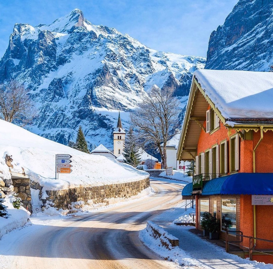 Snow-covered village street in Grindelwald with chalet houses, church steeple, and dramatic alpine mountains