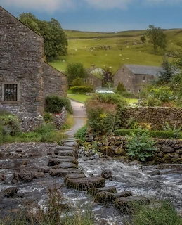 Stone cottages beside a stream with stepping stones in a rural countryside setting