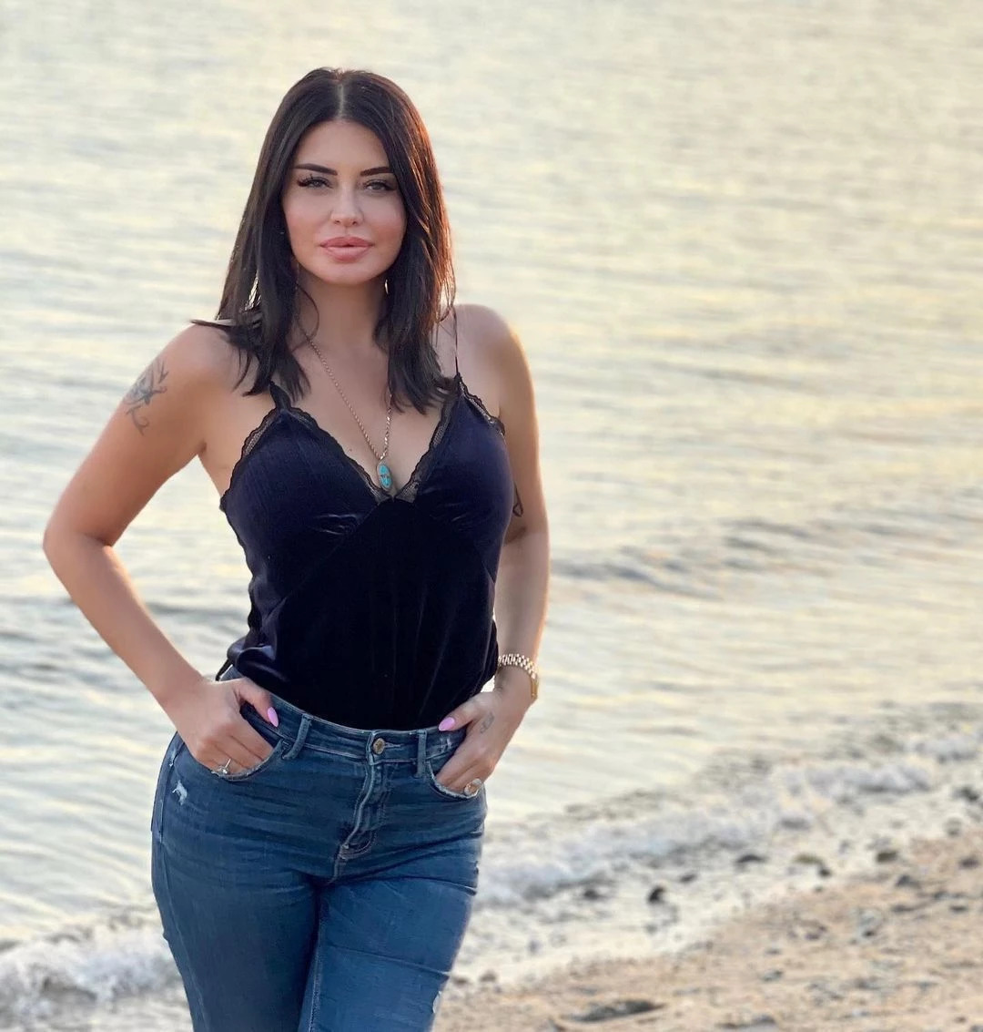 Woman in black top and jeans posing at beach with golden water behind her