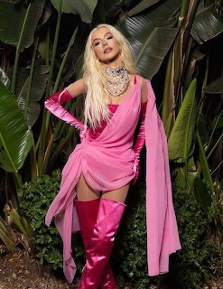 Blonde woman in pink satin dress, gloves, and boots posing in front of tropical plants