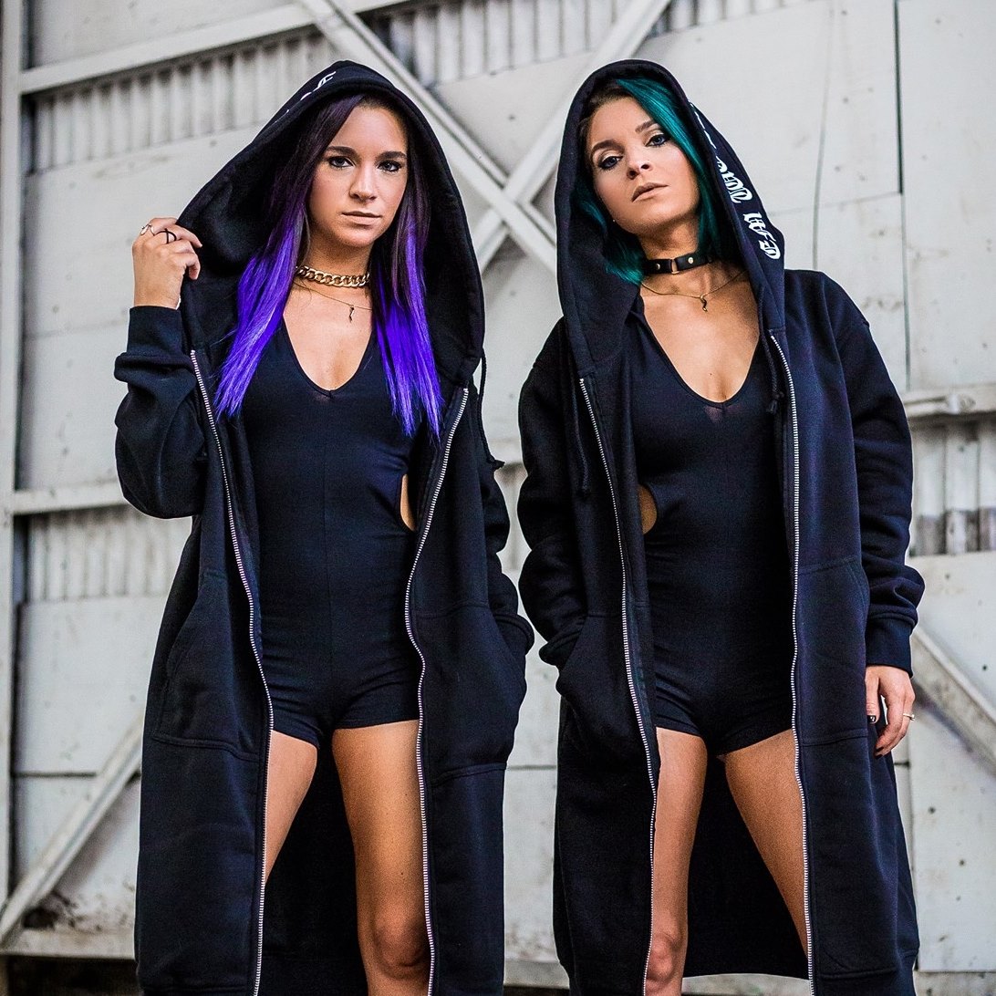 Two women in black hooded coats with purple and green hair standing against corrugated metal wall