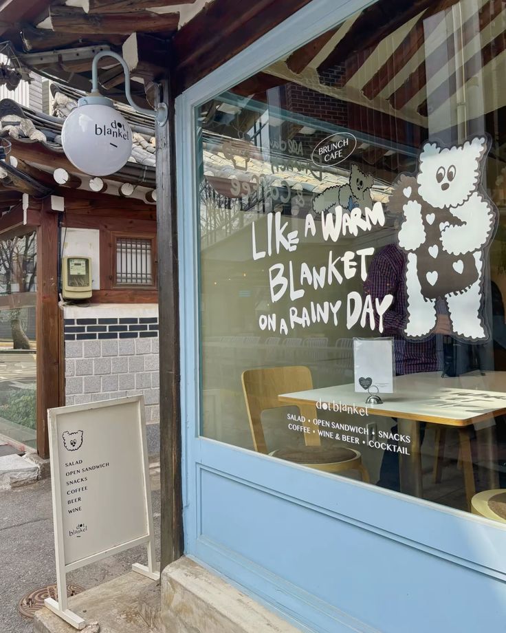 Korean cafe exterior with large window, hand-lettered quote and bear illustration, blue-framed door and wooden signboard