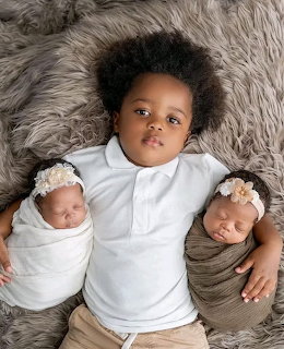 Young child lying on gray fur blanket between two swaddled newborn babies with floral headbands