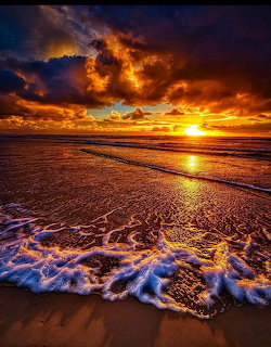 Vibrant sunset over ocean beach with dramatic clouds, golden reflections on wet sand, and white foam from incoming waves