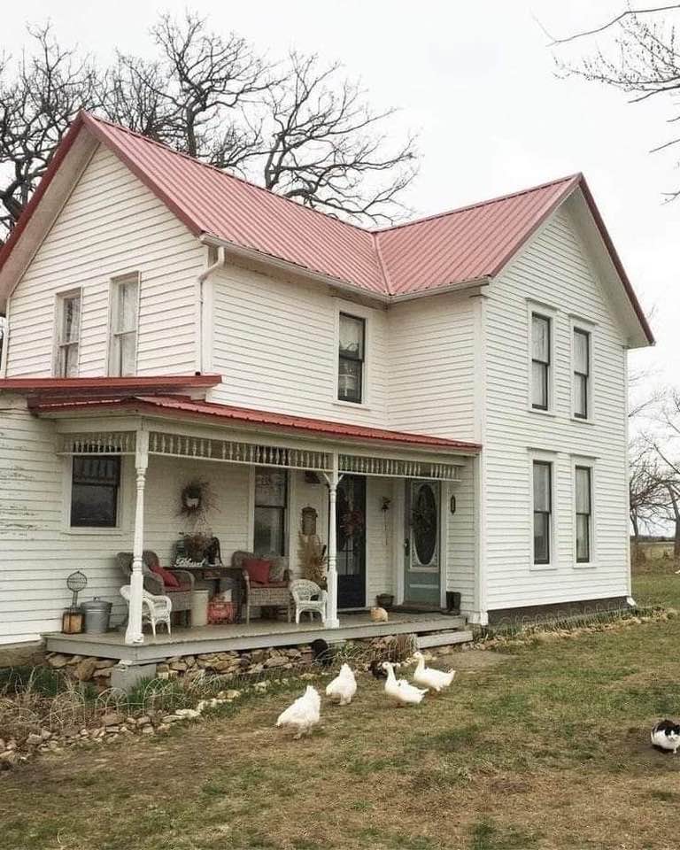 White two-story farmhouse with red metal roof and front porch, with geese roaming the yard