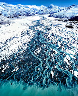 Aerial view of braided blue river channels flowing through glacial valley into turquoise lake with snowy mountains
