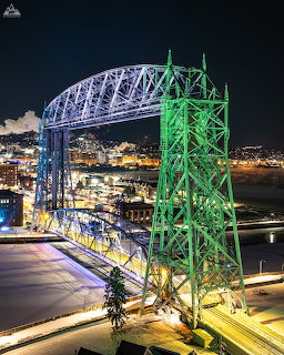 Duluth Aerial Lift Bridge lit up in blue and green at night over water with city lights behind