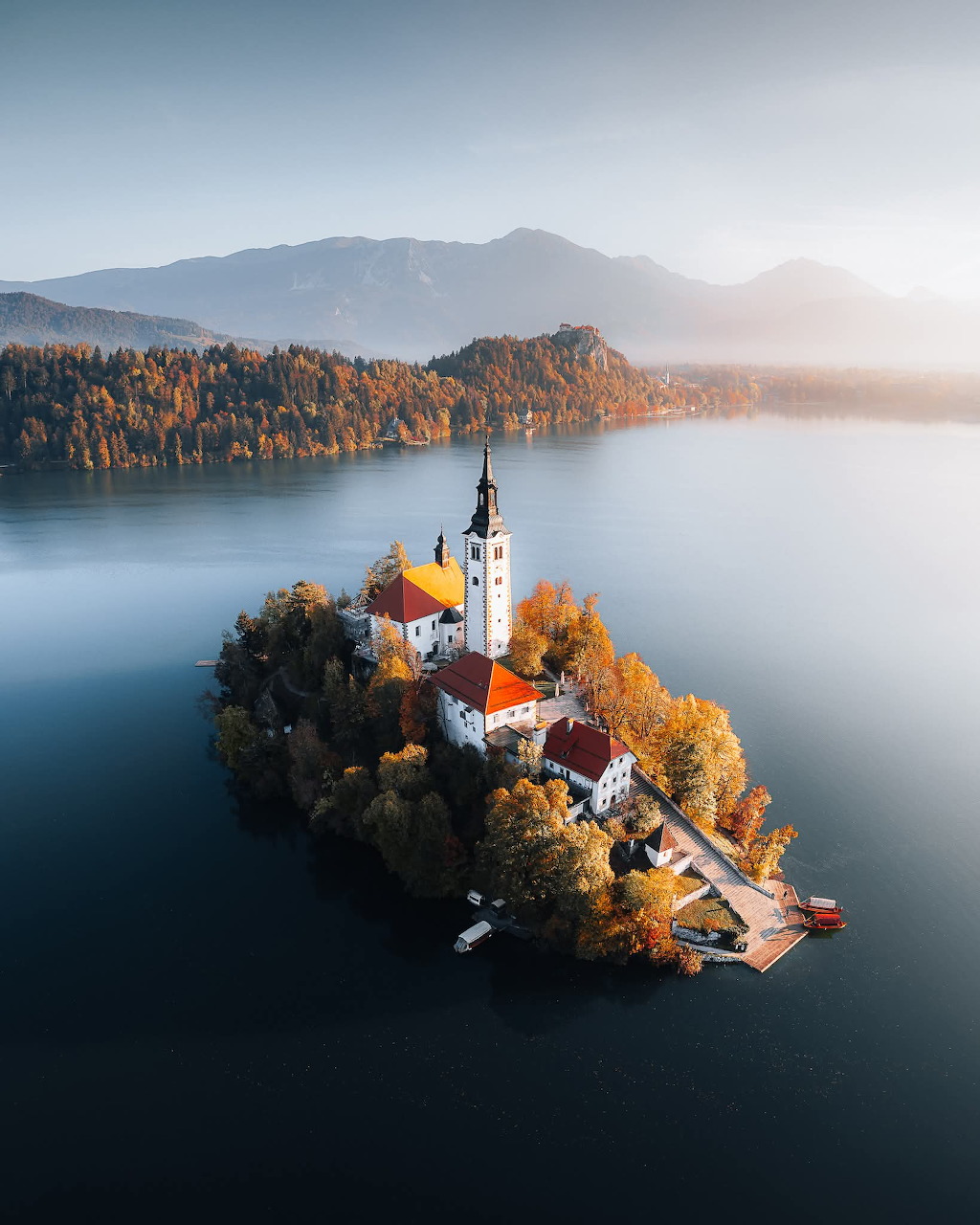 Aerial view of Lake Bled island with a white church tower and red roofs, surrounded by autumn trees and calm blue water