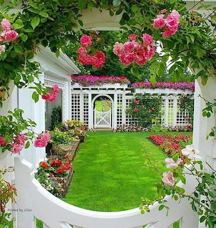 View through a rose-covered archway into a manicured garden with green lawn and white lattice gate