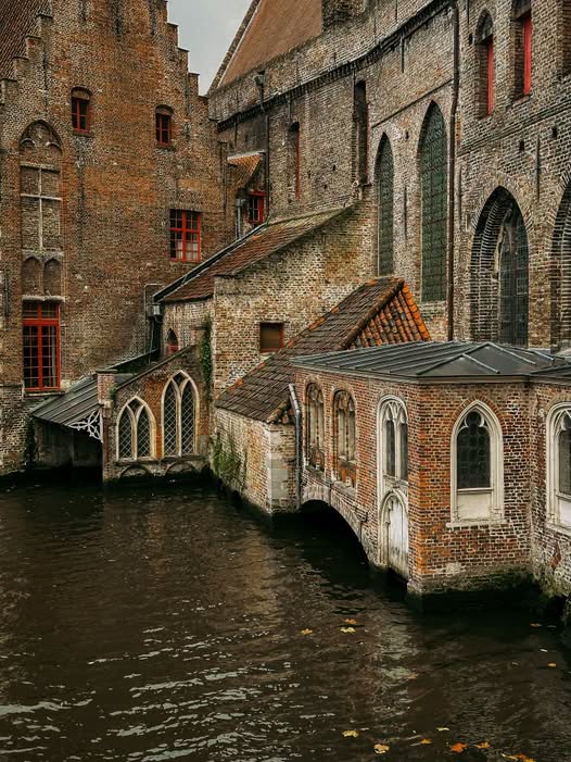 Historic brick buildings beside a canal in Bruges with arched windows and dark water