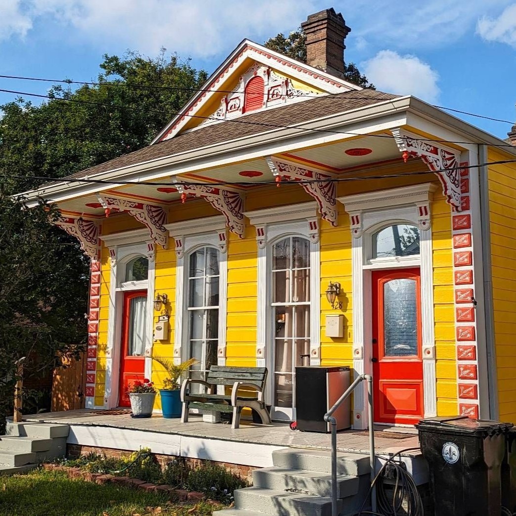Bright yellow shotgun house with red doors, decorative trim, and a covered porch under a blue sky