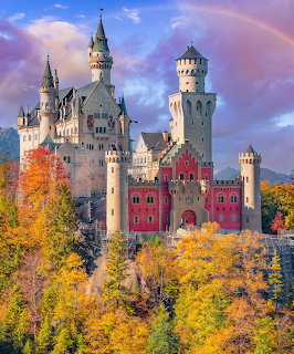 White castle with multiple towers surrounded by golden autumn trees beneath a rainbow and cloudy sky