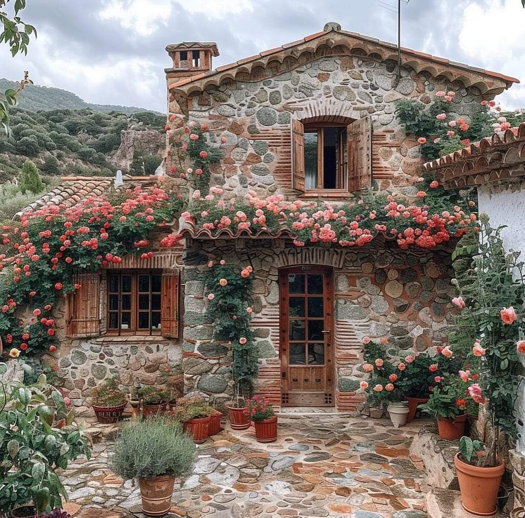 Rustic stone cottage with red tile roof, wooden door, window shutters, and abundant pink flowers in terracotta pots