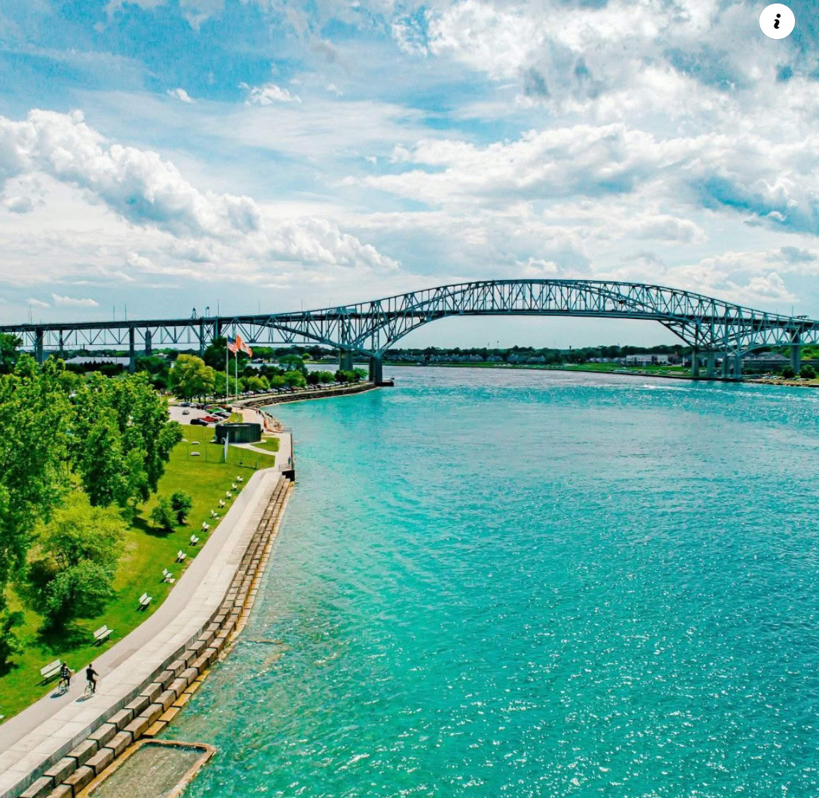 Steel arch bridge over blue water with a riverside path, trees, and cloudy sky