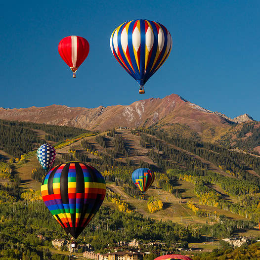 Several colorful hot air balloons flying over a mountain valley beneath a blue sky