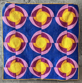 Geometric quilt with nine circular patterns in yellow, pink, and purple on blue background