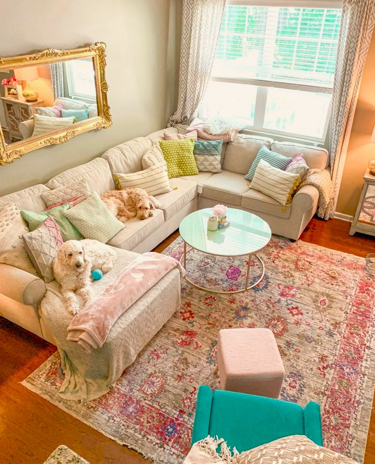 Bright living room with white sectional sofa, colorful pillows, two dogs, floral rug, and round glass coffee table