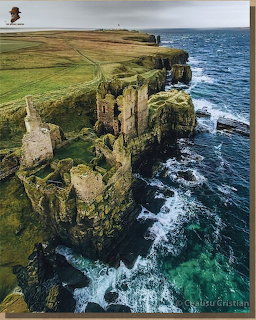 Aerial photo of a ruined Scottish castle on rocky sea cliffs with crashing waves and green fields