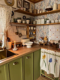 Vintage kitchen with green cabinets, wooden shelves, floral wallpaper, and rustic pottery displayed on open shelving