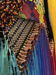 Colorful beaded textile art with geometric patterns, metallic cones, and rainbow fringe threads