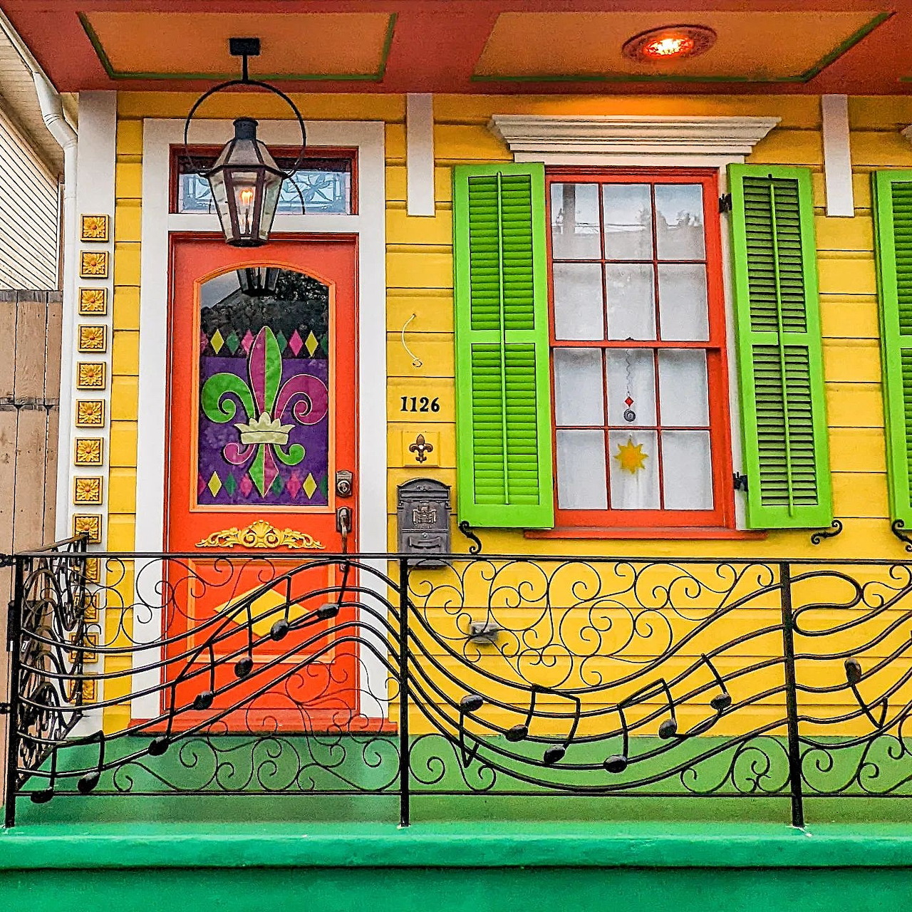 Bright porch with fleur-de-lis decorated door, green shutters, orange trim, yellow walls, and musical note iron railing