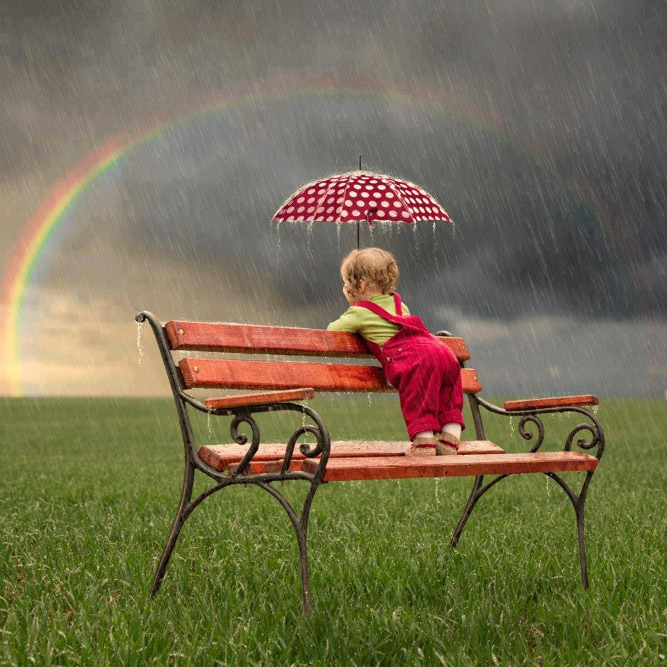 Child on red bench holding red polka dot umbrella in rain with rainbow in stormy sky