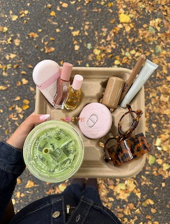 Hand holding a cardboard tray with makeup products, sunglasses, and a green iced drink on autumn pavement