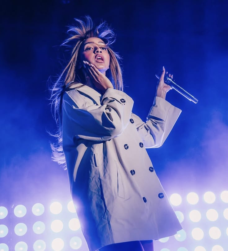 Woman in white coat singing on stage with blue stage lighting and spotlights behind her