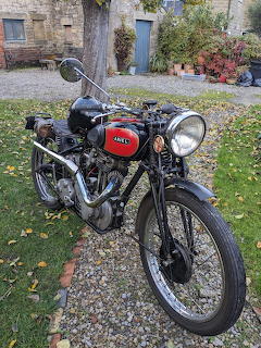 Vintage black and red Ariel motorcycle on gravel path with autumn foliage and stone building