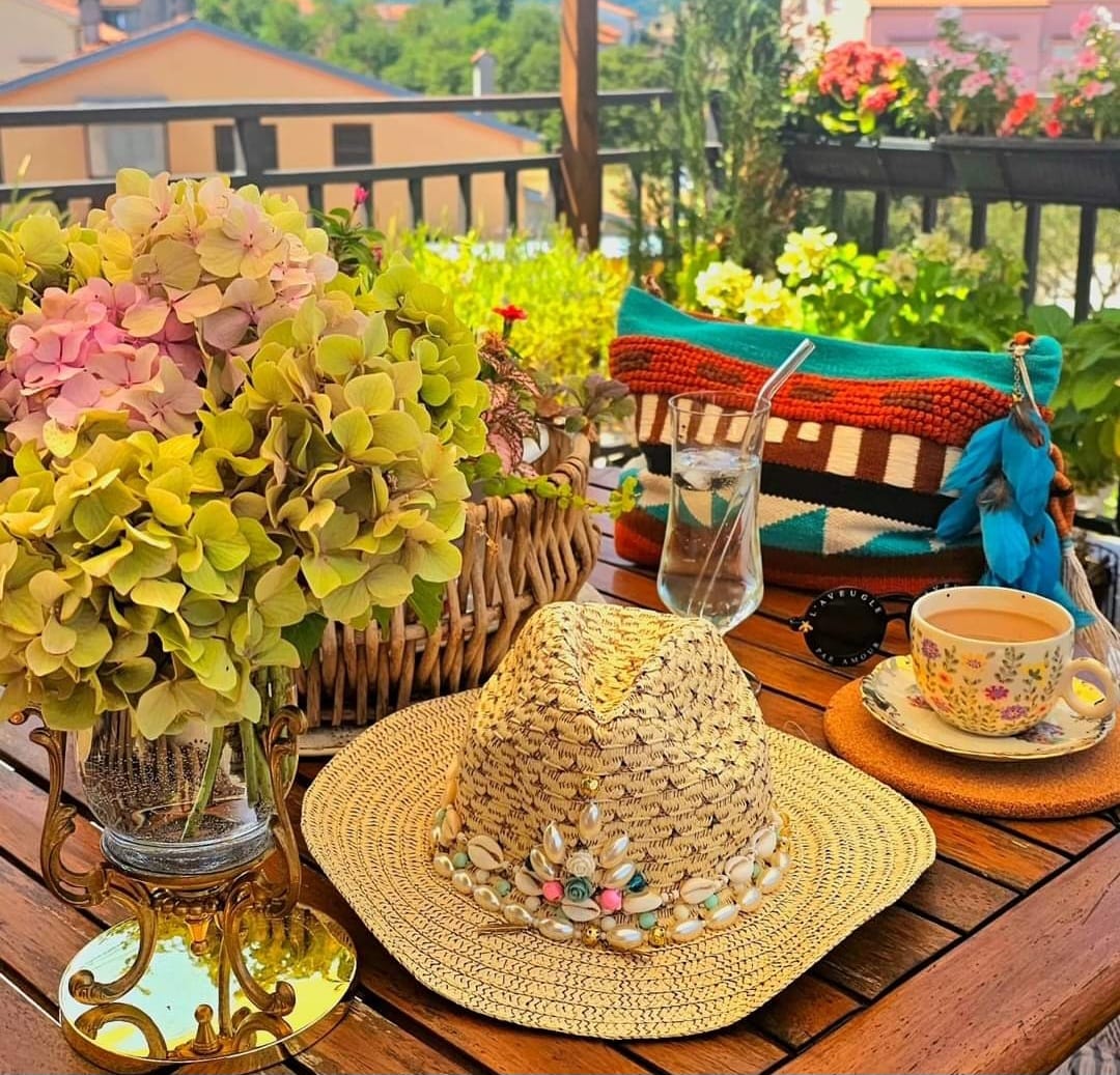 Woven straw hat with floral band on wooden balcony table next to teacup and hydrangea flowers