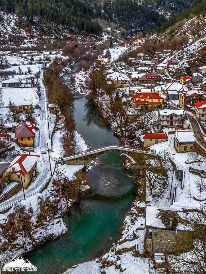 Aerial winter view of snow-covered Zagorohoria village with turquoise river and stone bridge, Greece