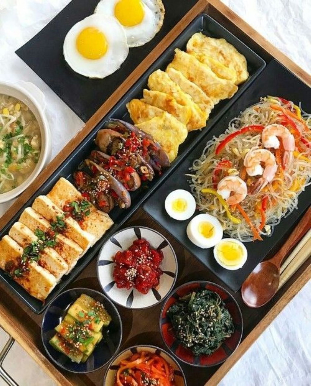 Overhead view of Korean meal with eggs, rolled omelets, meat skewers, seafood noodles, and side dishes on black plates