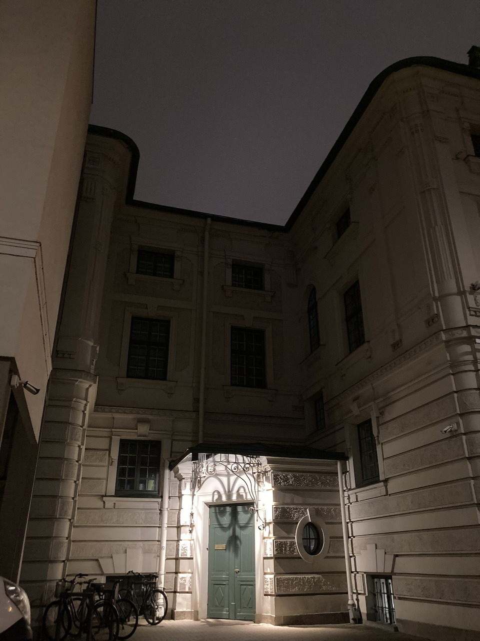 Night view of a stone building courtyard with an arched green door and bicycles by the entrance.