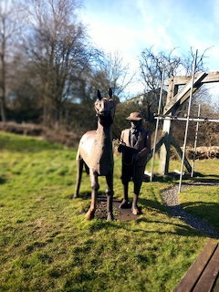 Person in dark clothing standing next to a white horse on grass with bare trees and wooden fence behind