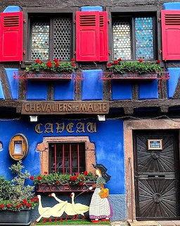 Colorful blue half-timbered building with red shutters, stained glass windows, and Chevaliers de Malte sign