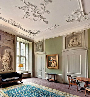 Ornate historic room with stucco ceiling decorations, classical reliefs, period furniture, and sage green walls