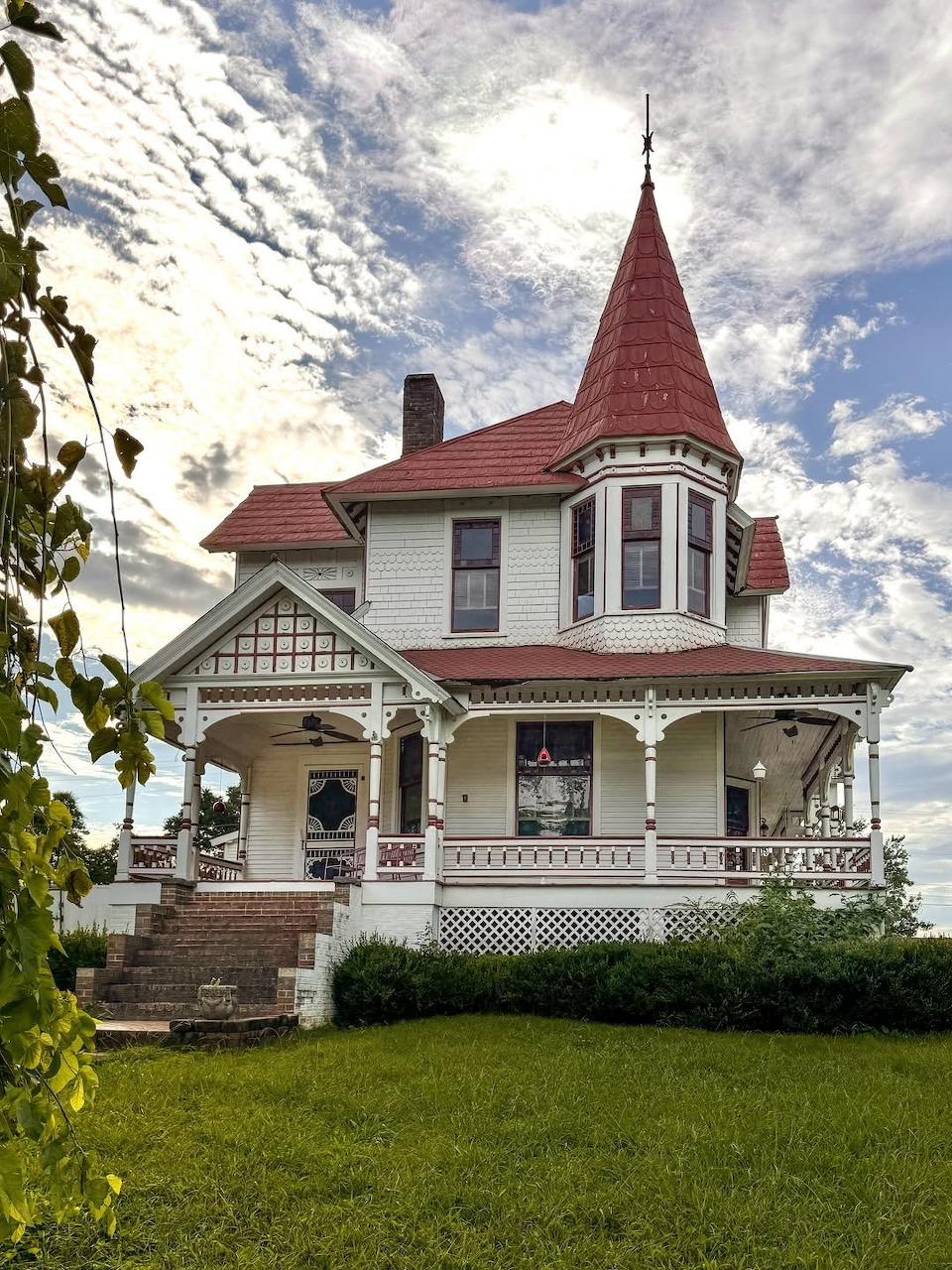 White Victorian house with red pointed turet roof, wraparound porch, and green lawn under cloudy sky