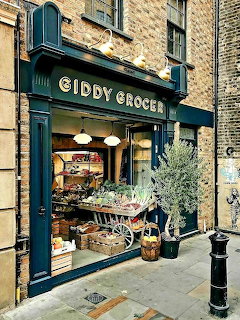Old-fashioned grocery storefront with fruit displays, potted plants, and dark teal trim on a city sidewalk